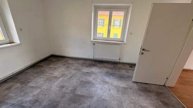 Leeres Zimmer mit grauem Laminatboden, weißen Wänden und zwei Fenstern, die viel Licht hereinlassen.