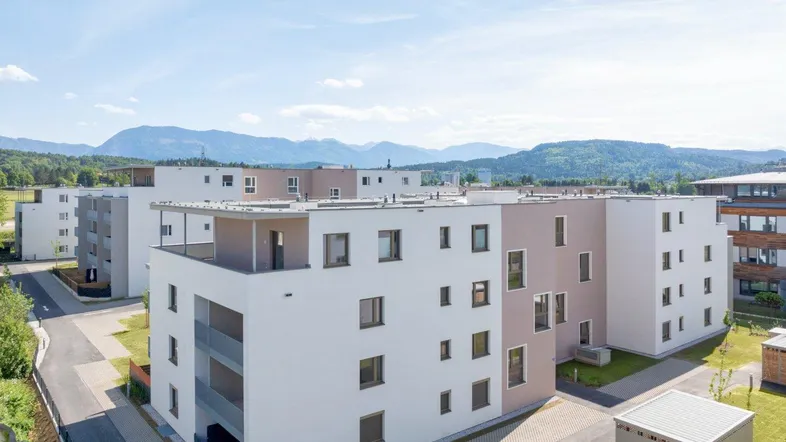 Moderne Wohngebäude in einer Siedlung mit Bergblick unter blauem Himmel und grüner Umgebung.