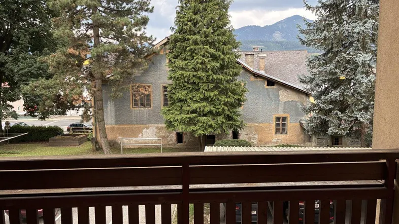 Blick vom Balkon auf ein älteres Gebäude mit Bäumen und Bergen im Hintergrund unter bewölktem Himmel.