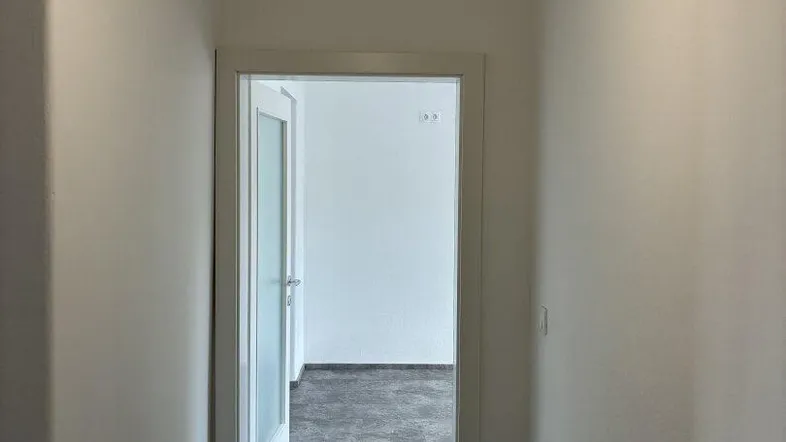 Schmaler Flur mit dunklem Boden und weiß gestrichenen Wänden, der zu einem weiteren Raum führt.