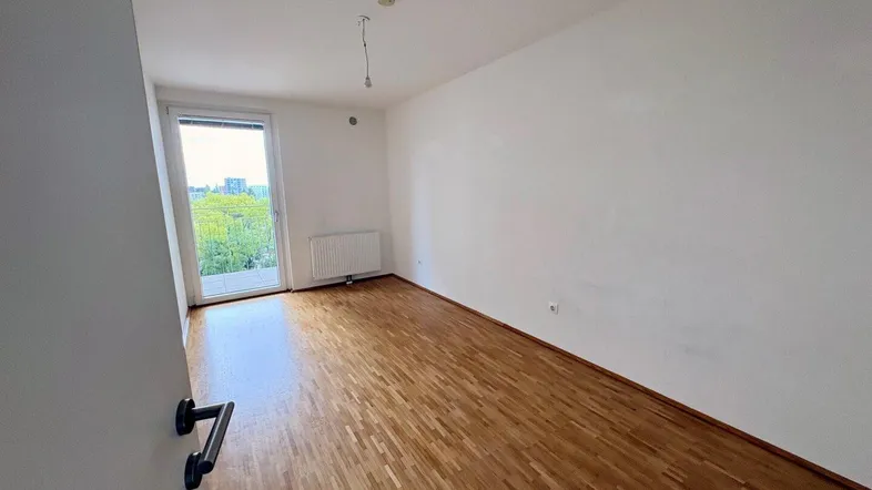 Helles Zimmer mit Parkettboden, weiß gestrichenen Wänden und Zugang zum Balkon.