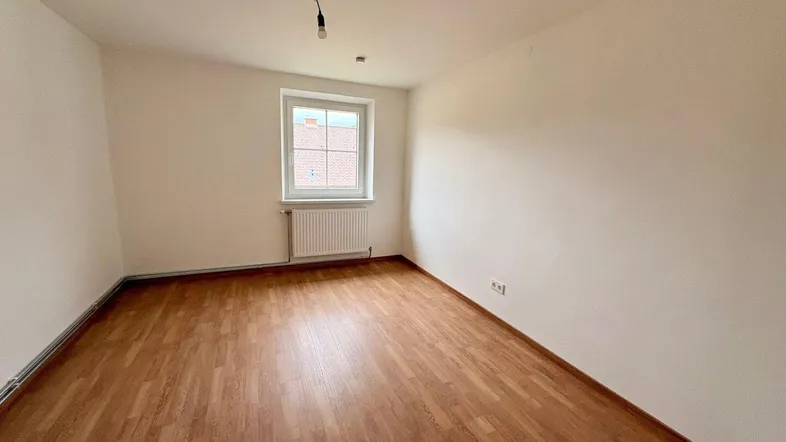 Leeres Zimmer mit Laminatboden, weißen Wänden und einem Fenster mit Heizkörper.