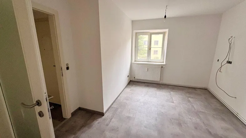 Helles, leeres Zimmer mit grauem Laminatboden und einem Fenster, bereit zur individuellen Gestaltung.