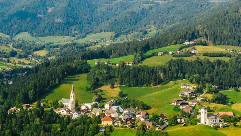 Panoramablick auf ein malerisches Dorf in einer grünen, hügeligen Landschaft mit Wäldern.