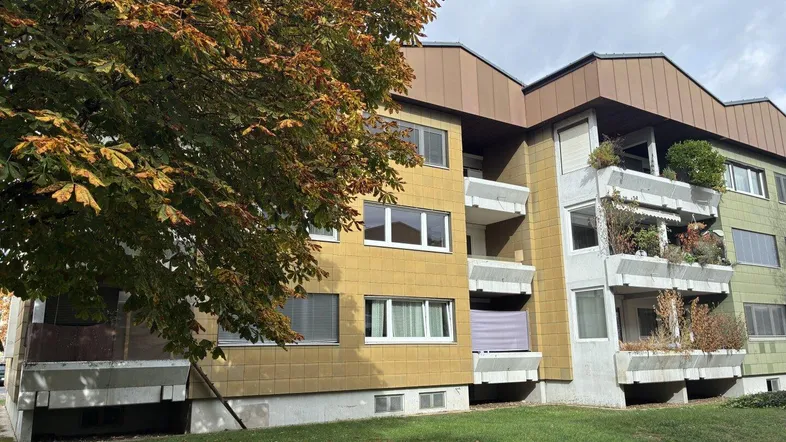 Mehrfamilienhaus mit gelber Fassade, Balkonen und herbstlicher Bepflanzung im Vordergrund.