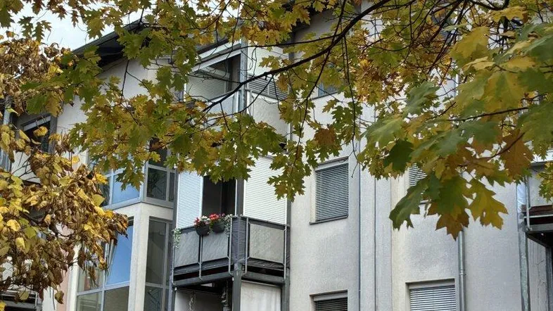 Mehrfamilienhausfassade mit Balkonen und herbstlichem Laub unter bewölktem Himmel.