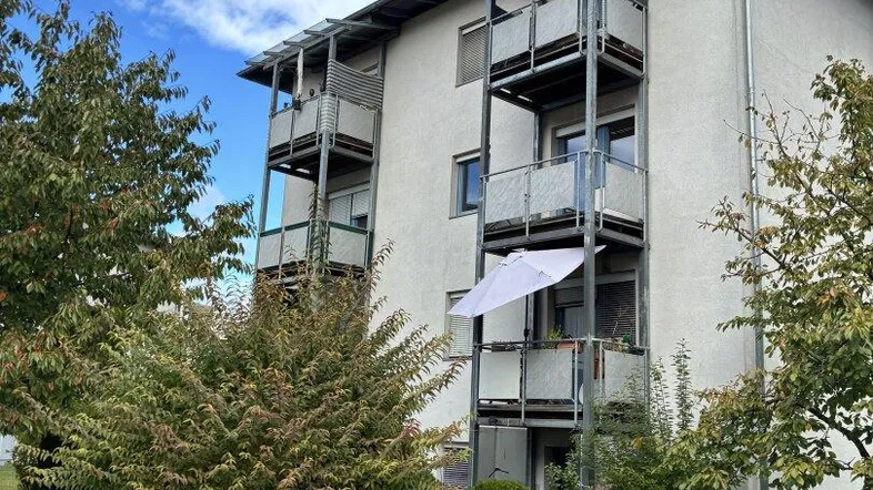 Mehrstöckiges Wohngebäude mit mehreren Balkonen und grüner Umgebung unter bewölktem Himmel.