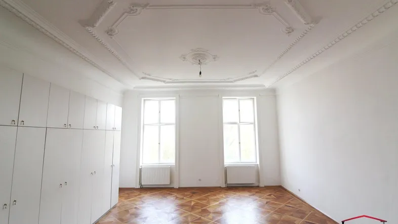 Geräumiges Zimmer mit elegantem Parkettboden, hohen Decken und dekorativem Stuck. Zwei große Fenster sorgen für viel Tageslicht.