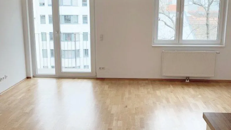 Helles, leeres Zimmer mit Parkettboden, weißen Wänden und Zugang zu einem Balkon.