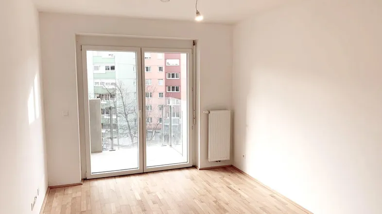 Helles Zimmer mit Parkettboden und Zugang zu einem Balkon, bietet Blick auf umliegende Gebäude.