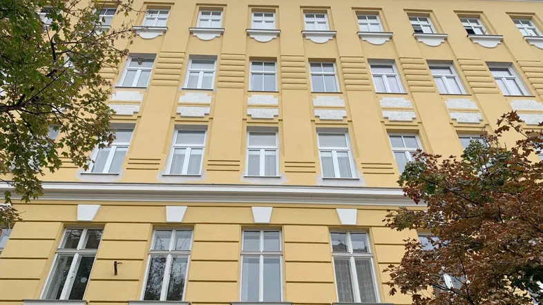 Gelbe Fassade eines Mehrfamilienhauses mit vielen Fenstern und dekorativen Elementen unter einem bewölkten Himmel.