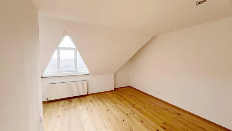 Helles Zimmer mit Holzboden, weißen Wänden und einem einzigartigen dreieckigen Fenster im Dachgeschoss.