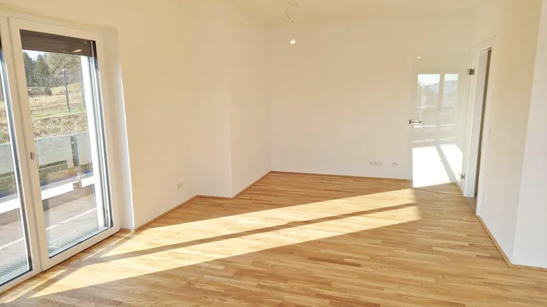 Helles, leeres Zimmer mit Parkettboden und Zugang zu einem Balkon oder einer Terrasse.