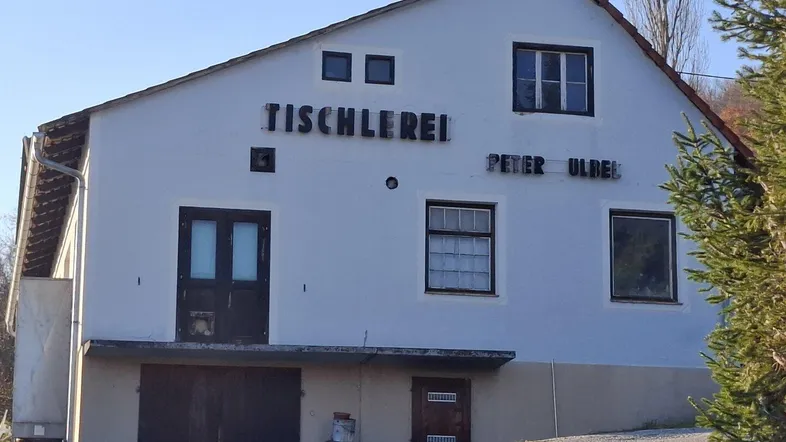 Das Gebäude zeigt eine weiße Fassade mit braunem Dach und Fenstern, sowie Beschriftungen einer ehemaligen Tischlerei.