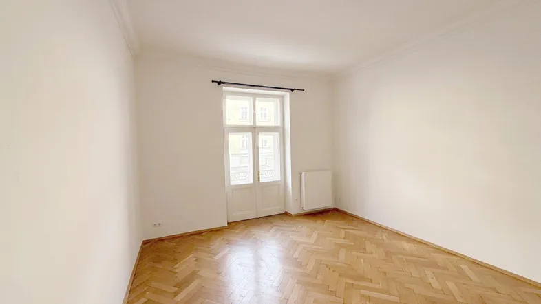 Helles Zimmer mit Fischgrätparkett, hohen Decken und Stuckelementen an der Decke.