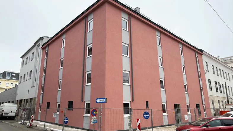Gebäudeaußenansicht mit rötlicher Fassade und grauen Fensterrahmen, umgeben von Bauzäunen.