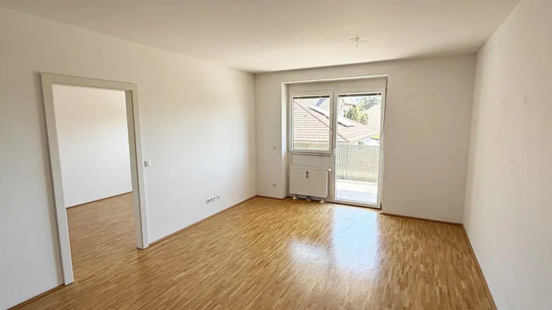 Helles Wohnzimmer mit Parkettboden, weißen Wänden und Zugang zu einem Balkon.