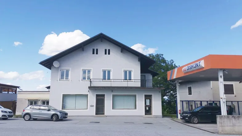 Helles Gebäude mit Satteldach, Balkon und Parkplätzen, angrenzend an eine Tankstelle.
