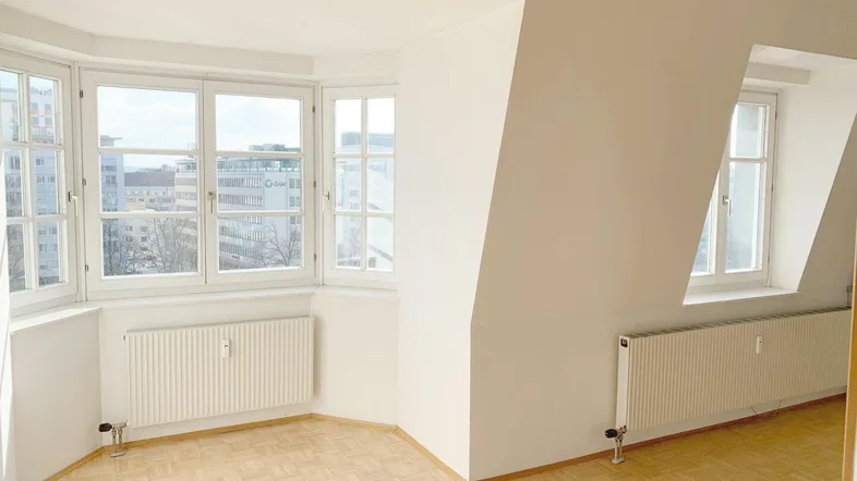 Helles Zimmer mit Parkettboden, großen Fenstern und schrägen Wänden, ideal als Wohn- oder Schlafbereich.