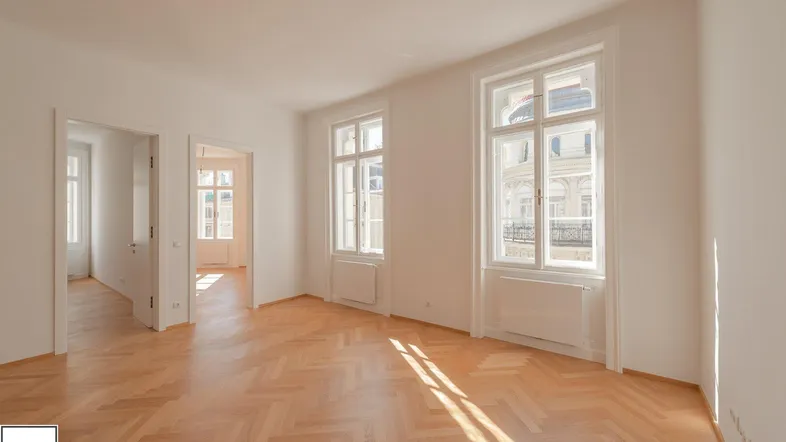Helles Zimmer mit Fischgrätparkett und hohen Fenstern, das viel Tageslicht hereinlässt.