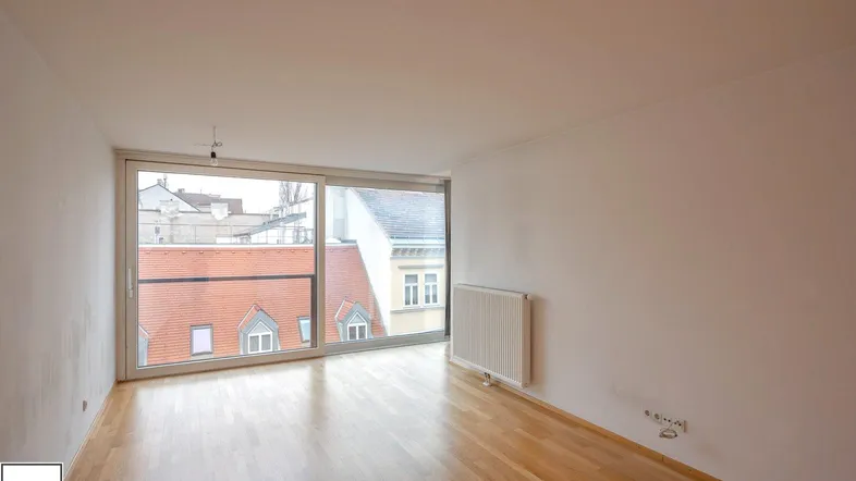 Helles Zimmer mit Holzboden und großen Fenstern, die einen Blick auf die umliegenden Dächer bieten.