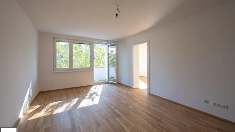 Helles, leeres Wohnzimmer mit Parkettboden und Zugang zu einem Balkon, durchflutet von Sonnenlicht.