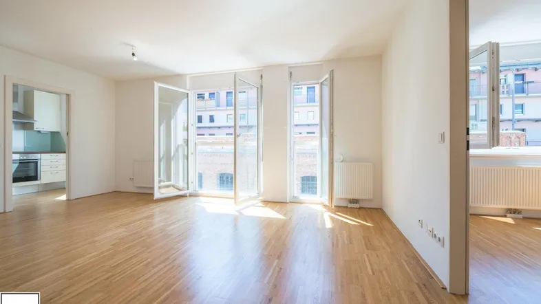 Helles, leeres Wohnzimmer mit Parkettboden und großen Fenstern, die viel Tageslicht hereinlassen.