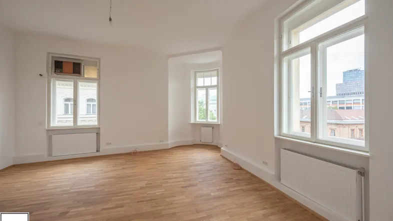 Heller, leerer Raum mit Parkettboden und mehreren Fenstern, die viel Tageslicht hereinlassen.