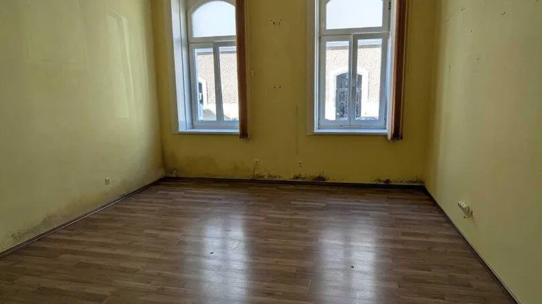 Leeres Zimmer mit gelben Wänden, Laminatboden und zwei hohen Bogenfenstern.