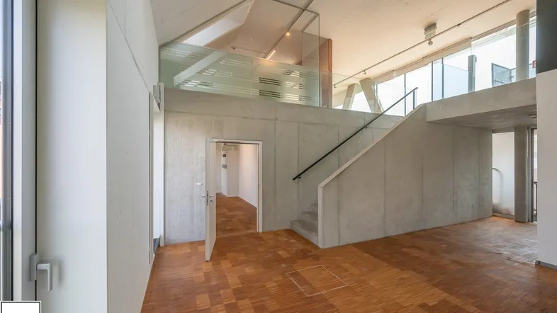 Geräumiger Innenraum mit Holzparkettboden, Betondecken und einer modernen Treppe mit Glasgeländer.