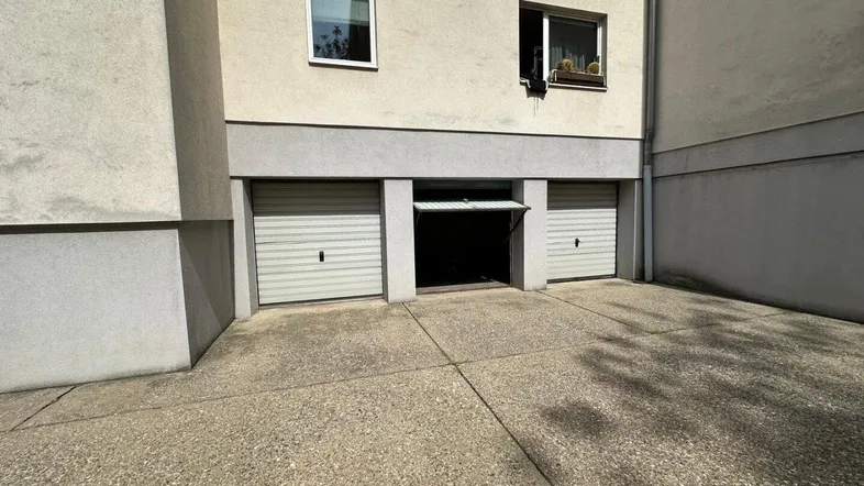 Mehrere Garagentore an der Gebäudefassade, eines davon leicht geöffnet, mit einem gepflasterten Vorplatz.