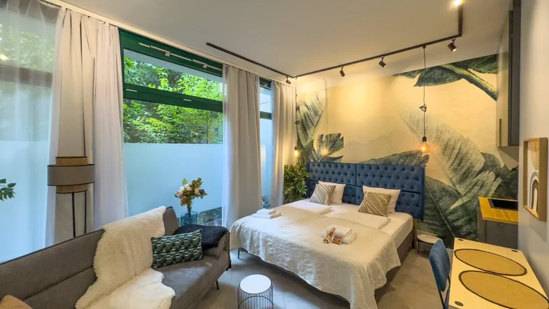 Modernes Schlafzimmer mit Doppelbett, Sofa und tropischer Wandtapete, hell durch große Fenster.