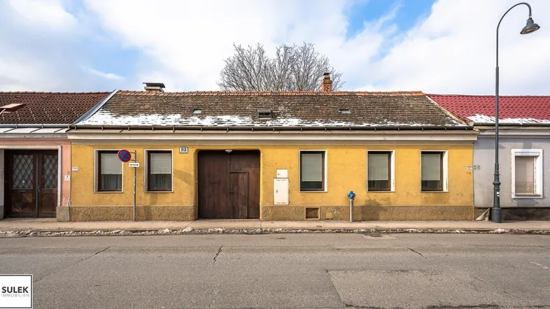 Gelbe Fassade eines älteren Gebäudes mit braunem Tor und Fenstern zur Straße hin.