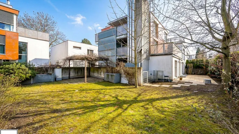 Sonniger Gartenbereich mit Rasenfläche, Bäumen und Blick auf die moderne Architektur des Gebäudes.
