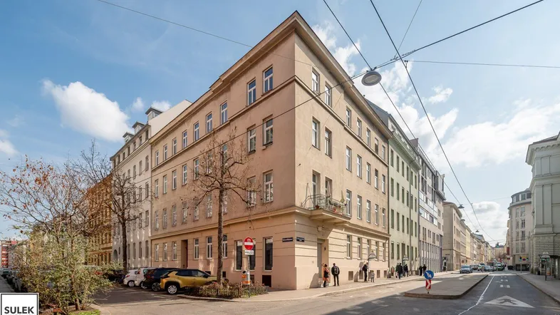 Ansicht des Eckgebäudes mit mehreren Stockwerken und parkenden Autos auf der Straße unter blauem Himmel.