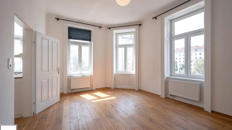 Helles Zimmer mit Holzboden und großen Fenstern, das viel Tageslicht hereinlässt.