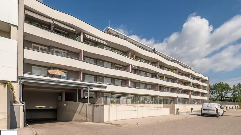 Modernes Mehrfamilienhaus mit geschwungener Fassade, vielen Balkonen und Tiefgaragenzufahrt.