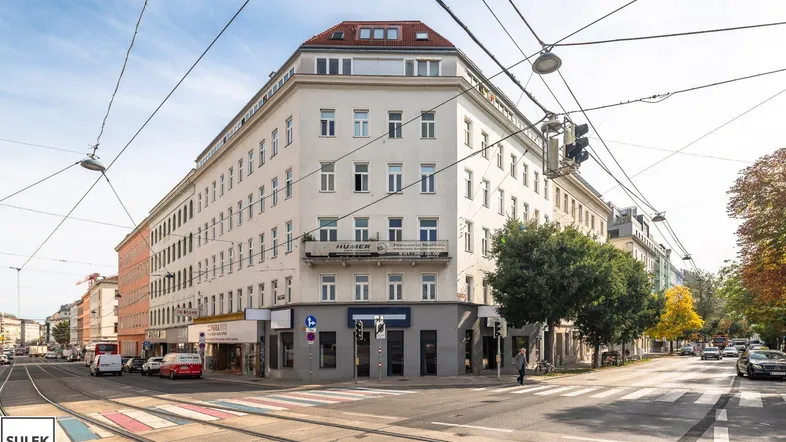 Großes Eckgebäude an einer belebten Straße mit Straßenbahnschienen und Geschäften im Erdgeschoss.
