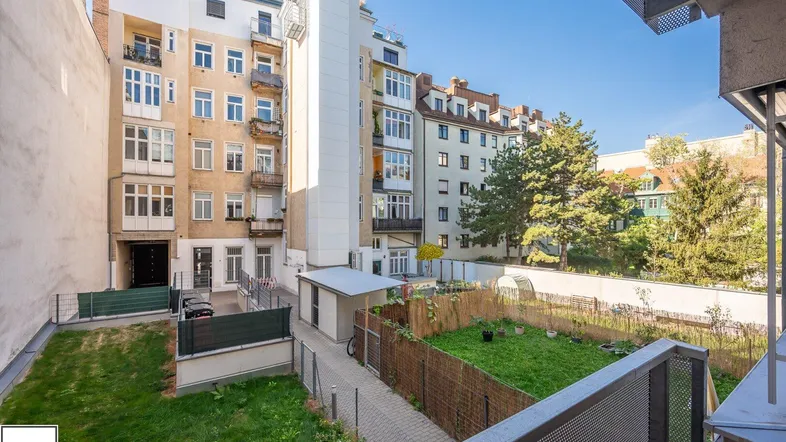 Blick von einem Balkon auf den gemeinschaftlichen Gartenbereich mit umliegenden Wohngebäuden unter blauem Himmel.