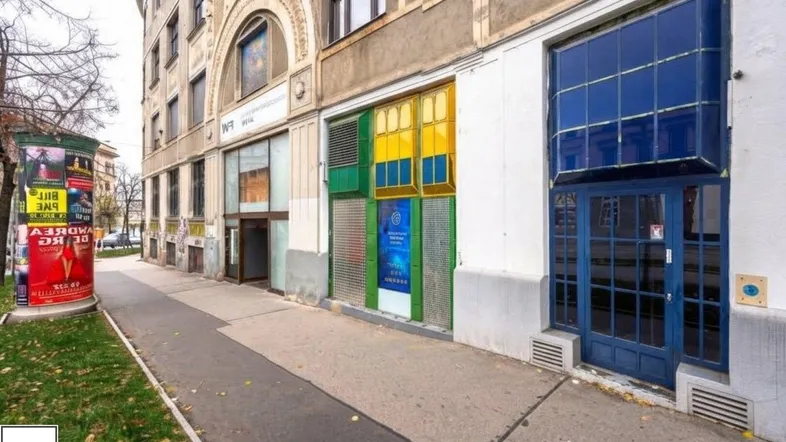 Eine belebte Straße mit einem historischen Gebäude und verschiedenen Ladenfronten im Erdgeschoss, umgeben von Bäumen.
