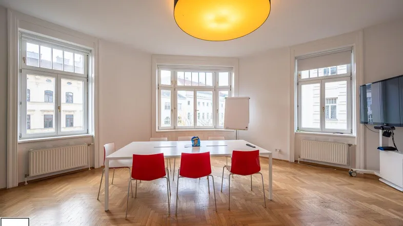 Geräumiger Büroraum mit Fischgrätparkett, großen Fenstern und einem Konferenztisch mit roten Stühlen.