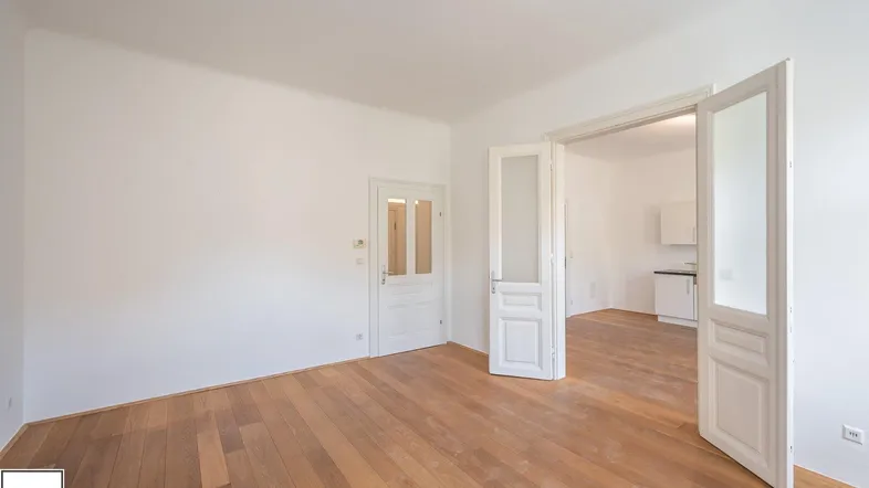 Helles Zimmer mit Holzboden und weißen Wänden, Türen führen in einen weiteren Raum.