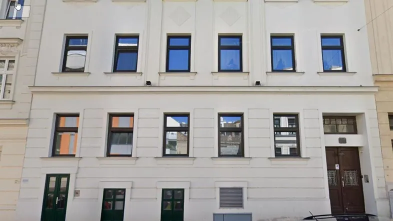 Die weiße Fassade des Mehrfamilienhauses mit mehreren Fenstern und einem Eingangsbereich an einer Stadtstraße.