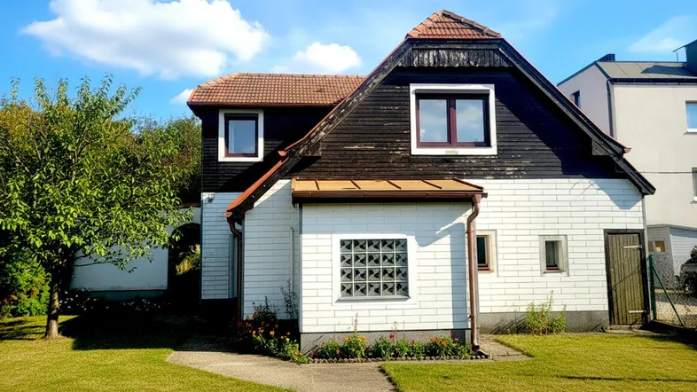 Hausfassade mit weißer und dunkler Holzverkleidung, umgeben von einem gepflegten Rasen und Bäumen.