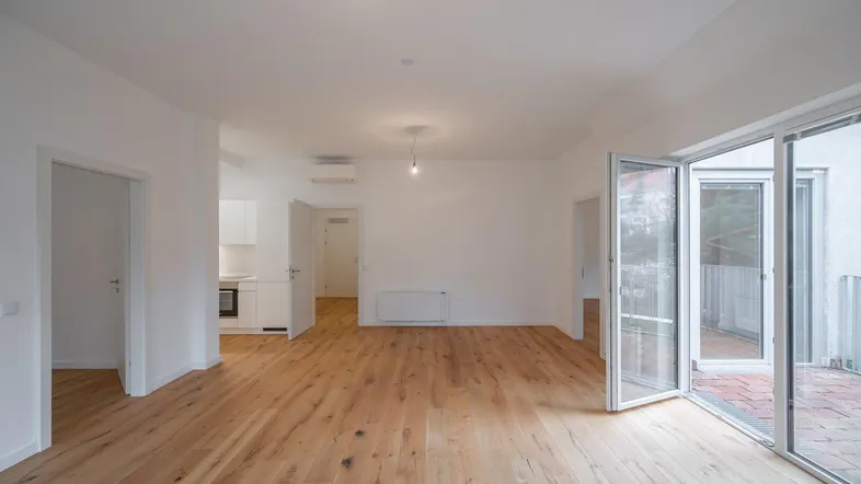 Helles, leeres Wohnzimmer mit Holzboden und Zugang zu einer Terrasse und offener Küche.