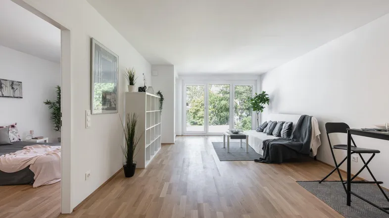 Helles Wohnzimmer mit Parkettboden, großen Fenstern und Blick ins Grüne, angrenzend an ein Schlafzimmer.