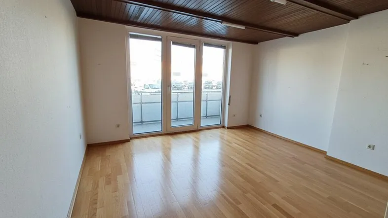 Heller, leerer Raum mit Parkettboden, Holzbalkendecke und Zugang zu einem Balkon.