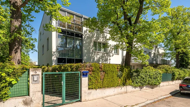 Modernes Mehrfamilienhaus mit großen Fenstern, umgeben von viel Grün und Bäumen.