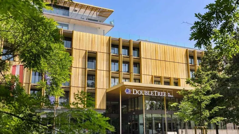 Moderne Hotelfassade mit Holzverkleidung und großen Fenstern, umgeben von grünen Bäumen unter blauem Himmel.