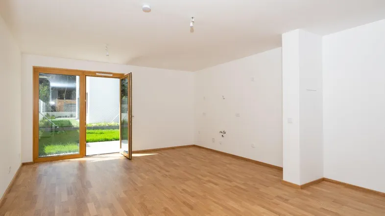Helles, leeres Zimmer mit Holzboden und Zugang zu einem kleinen Gartenbereich.